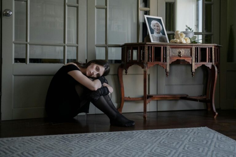 A solitary woman in black attire sits on the floor in mourning inside her home.