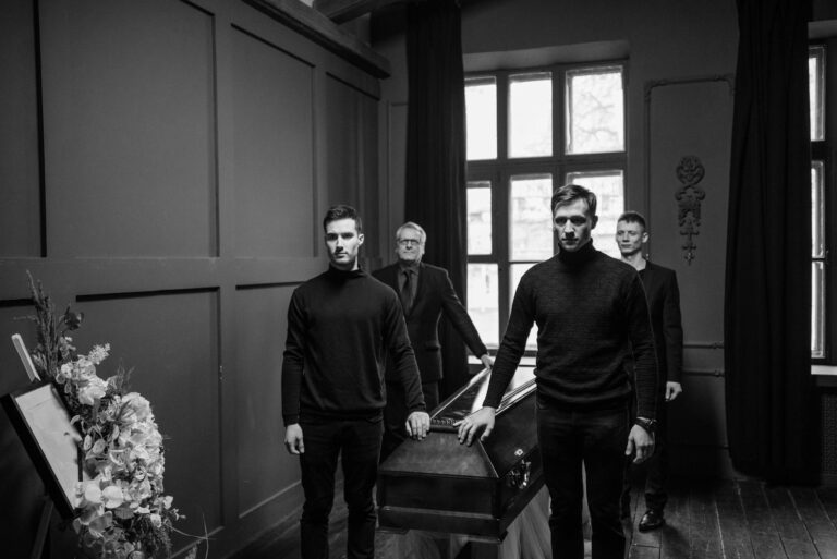Group of men in black attire solemnly carrying a coffin in an indoor setting.