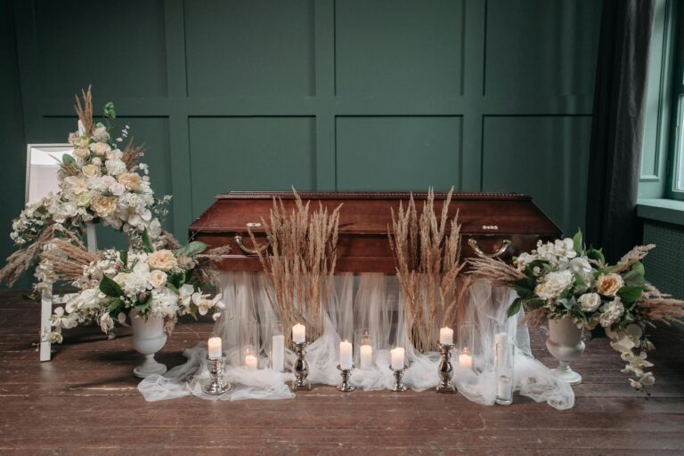 Wooden coffin surrounded by candles and floral arrangements in a serene funeral setting.