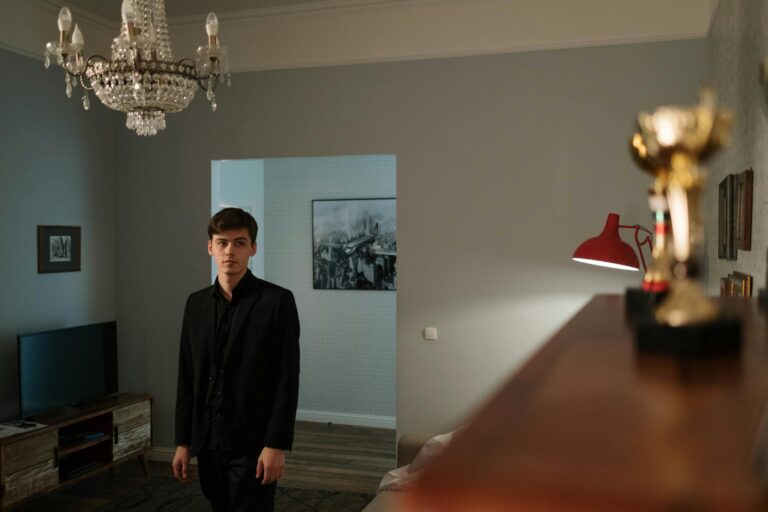 Young man in black suit stands in a classic living room with trophies and chandelier.